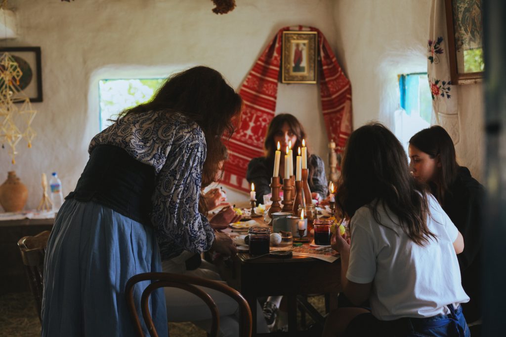 A group of people at the table decorating a traditional ukrainian easter egg - pysanka using wax and dyes. They sit in the old traditional countryside house with images and ritual embroidered linen cloths.. A woman in a blue dress leans over the table to help after showing a masterclass. Table is full of candles and the atmosphere is cozy