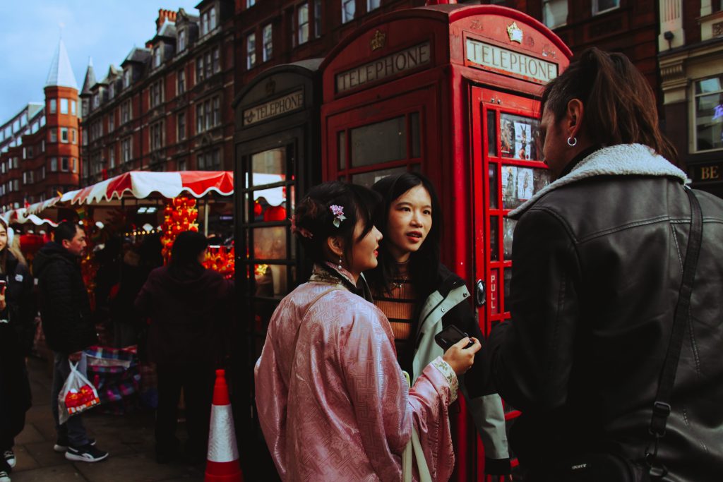 Three Asian people talk to each other on the streets of Chinatown during Chinese New Year celebration. One girl is wearing a traditional pink kimono with a festive chinese hairstyle. London phone booths are on the background