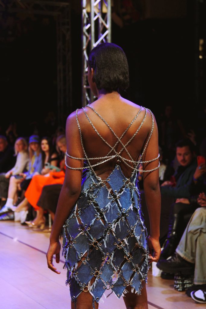 A black model presenting a dress at the catwalk at London Fashion Week 2024. It is made of small diamond shaped denim pieces that are connected with chains creating a beautiful pattern on her back. The audience on the background is watching her