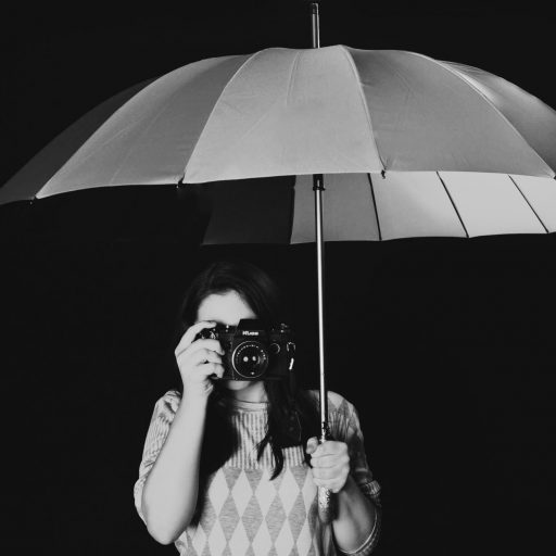 Black and white photo of a girl holding a stried umbrella and a camera to cover her face